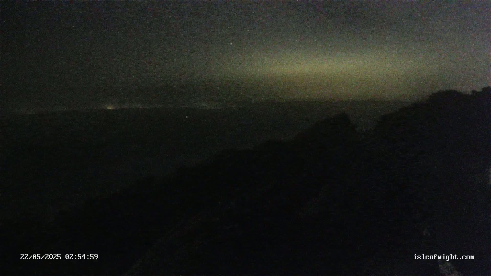 A dark, nighttime view shows a distant horizon line with a faintly lit sky above, partially obscured by a silhouetted foreground of trees or hills.