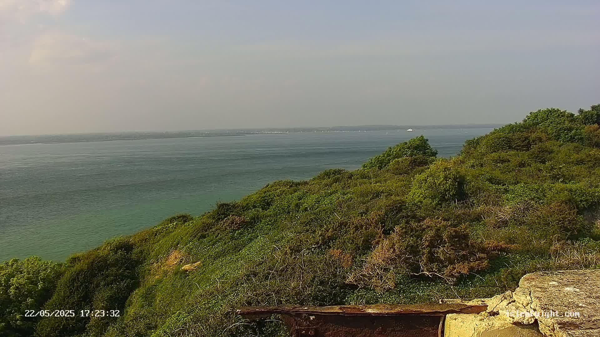 A calm, hazy day reveals a body of water extending to a distant shoreline, viewed from a verdant cliff top with some rusted metal in the foreground.