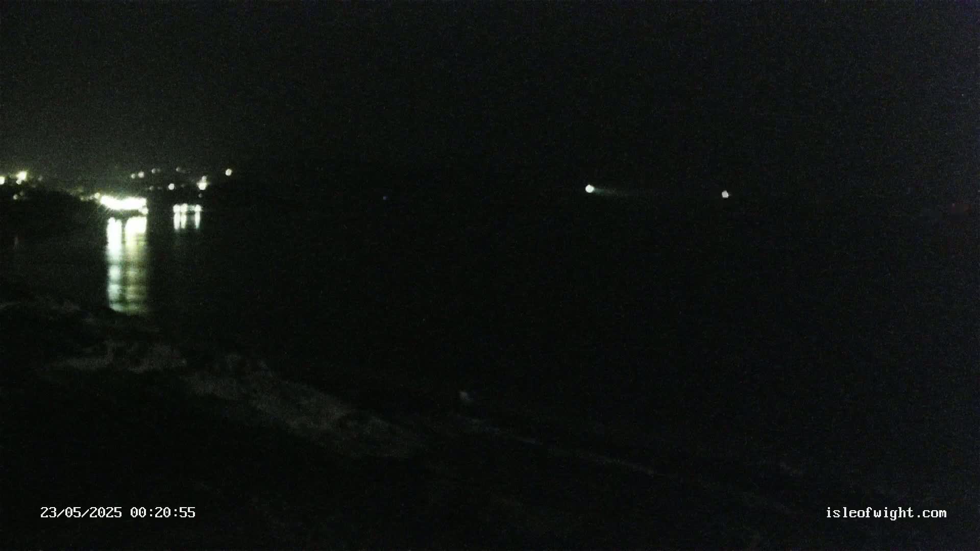 At night, a dark body of water reflects the lights of a distant town under a clear sky.