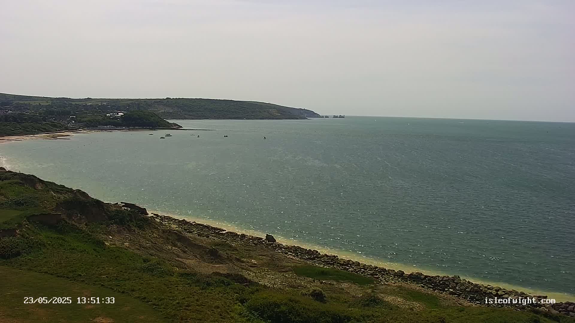 A coastal scene on a partly sunny day shows a calm sea stretching to a distant headland, with a sandy beach curving along a grassy cliff in the foreground.
