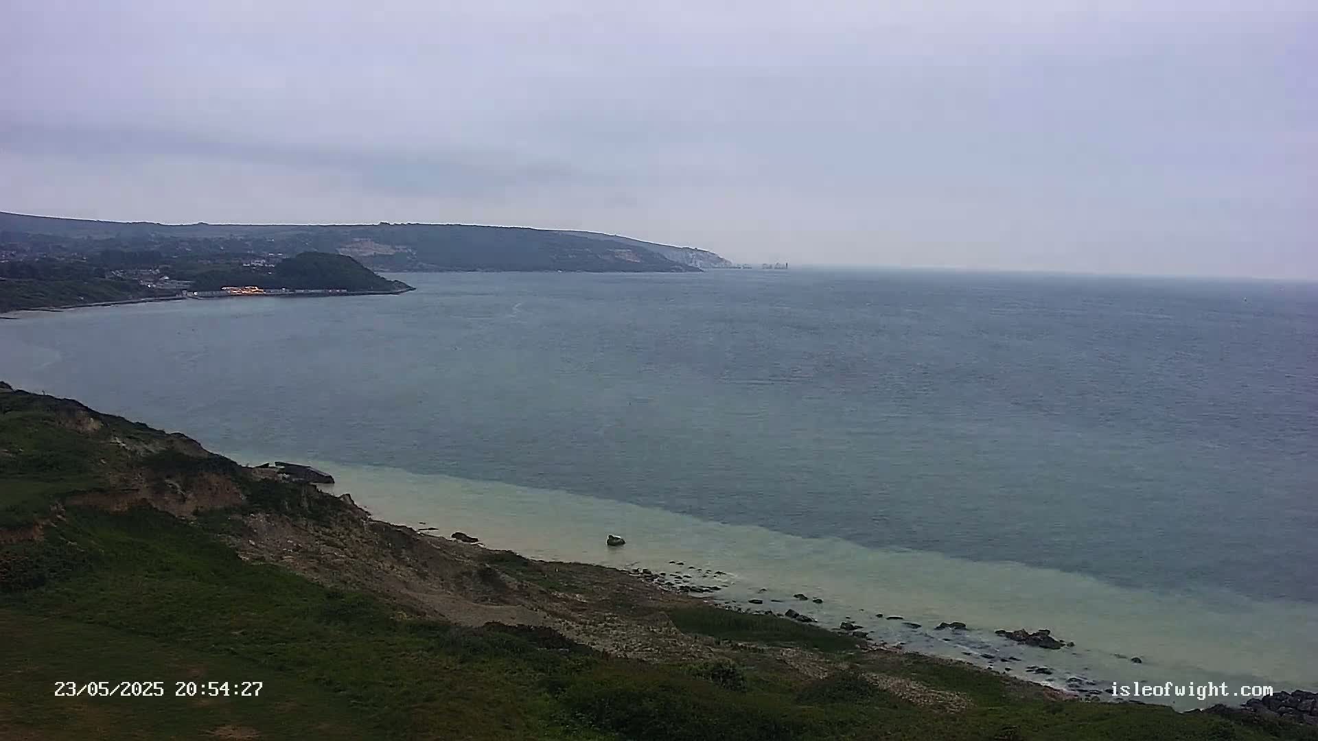 A coastal scene under an overcast sky shows a body of water meeting a grassy cliffside with a small beach area; the water appears calm.