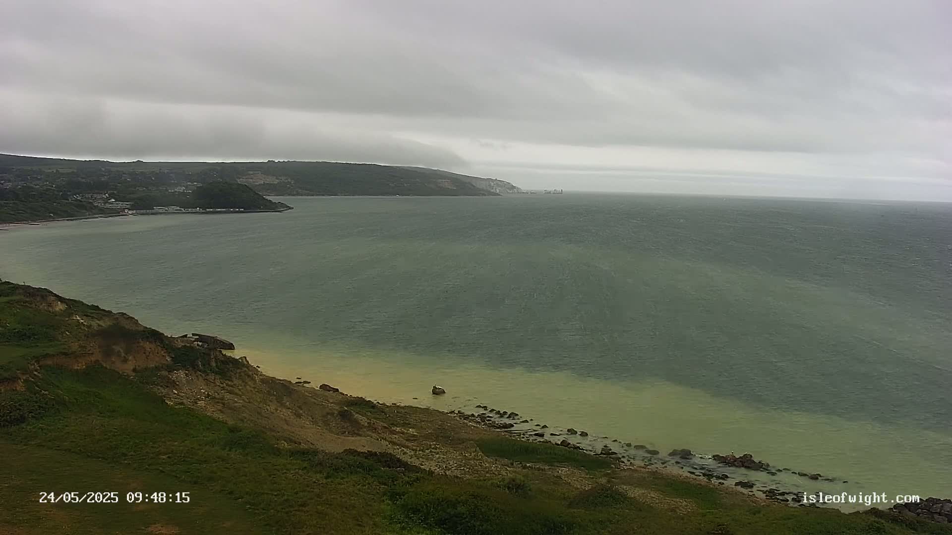 A coastal scene shows a gray, overcast sky over a green-tinged sea meeting a sandy beach at the base of a grassy cliff.
