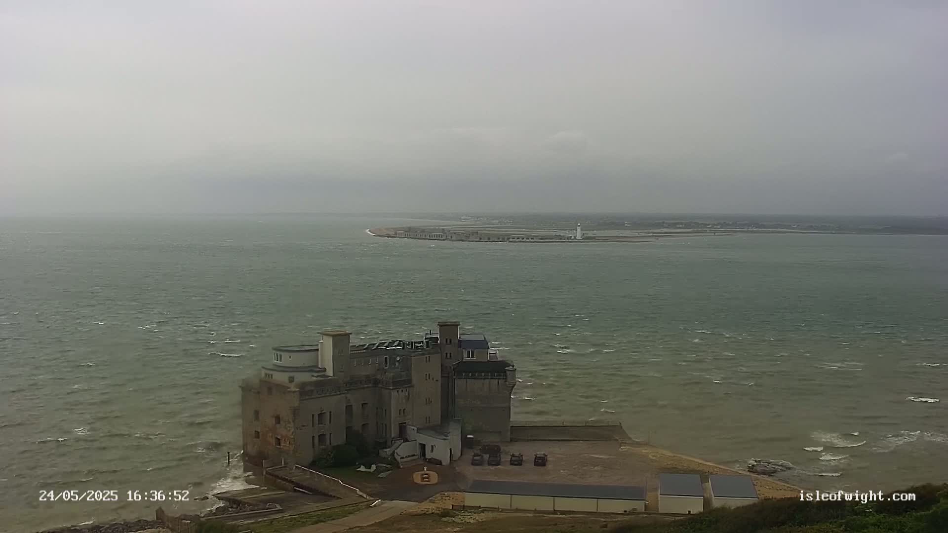 A stone building sits on a cliff overlooking a choppy sea, with a distant landmass and lighthouse visible under an overcast sky.