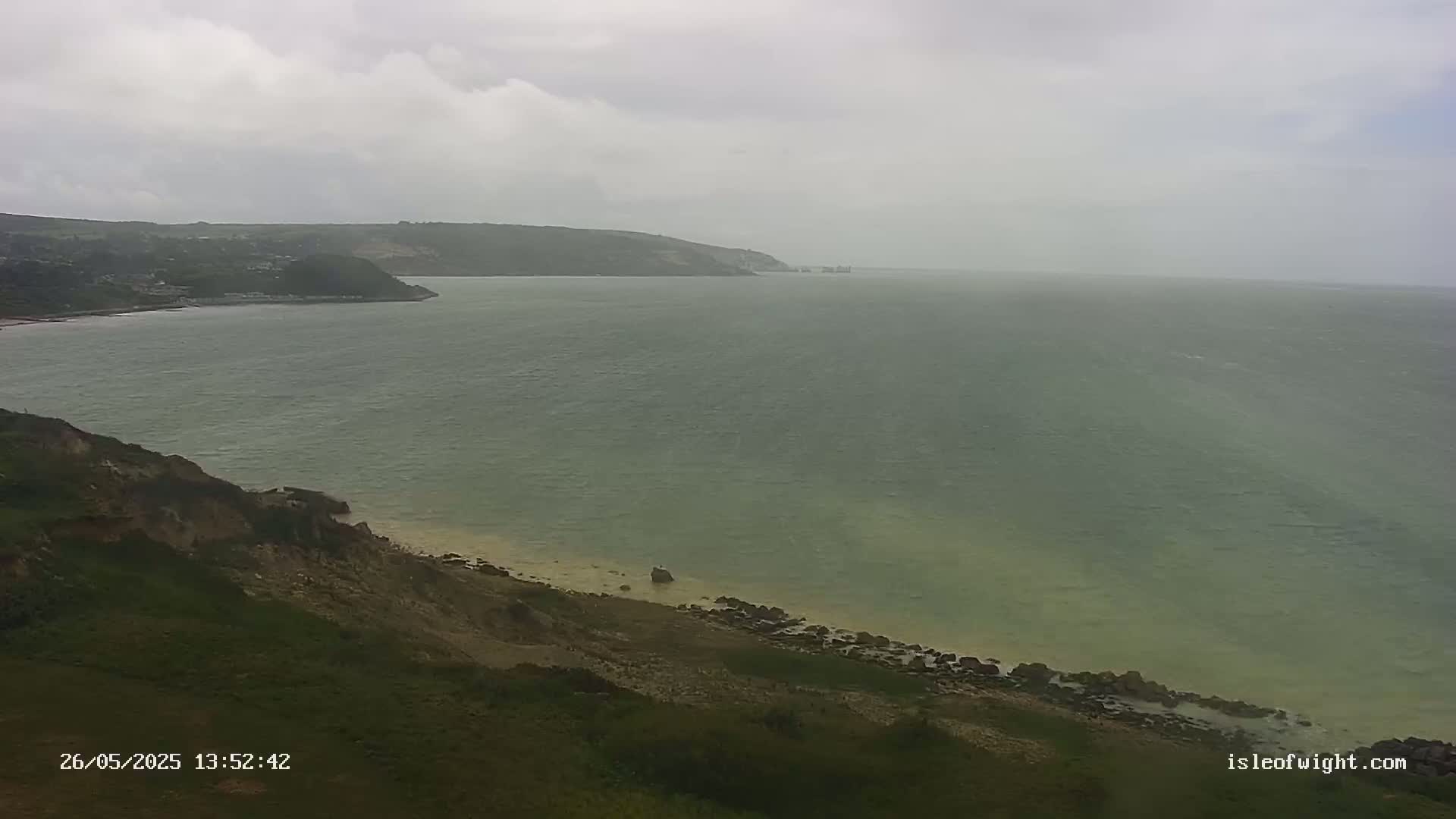 A coastal view shows a calm sea under an overcast sky, with green hills and a sandy beach in the foreground.