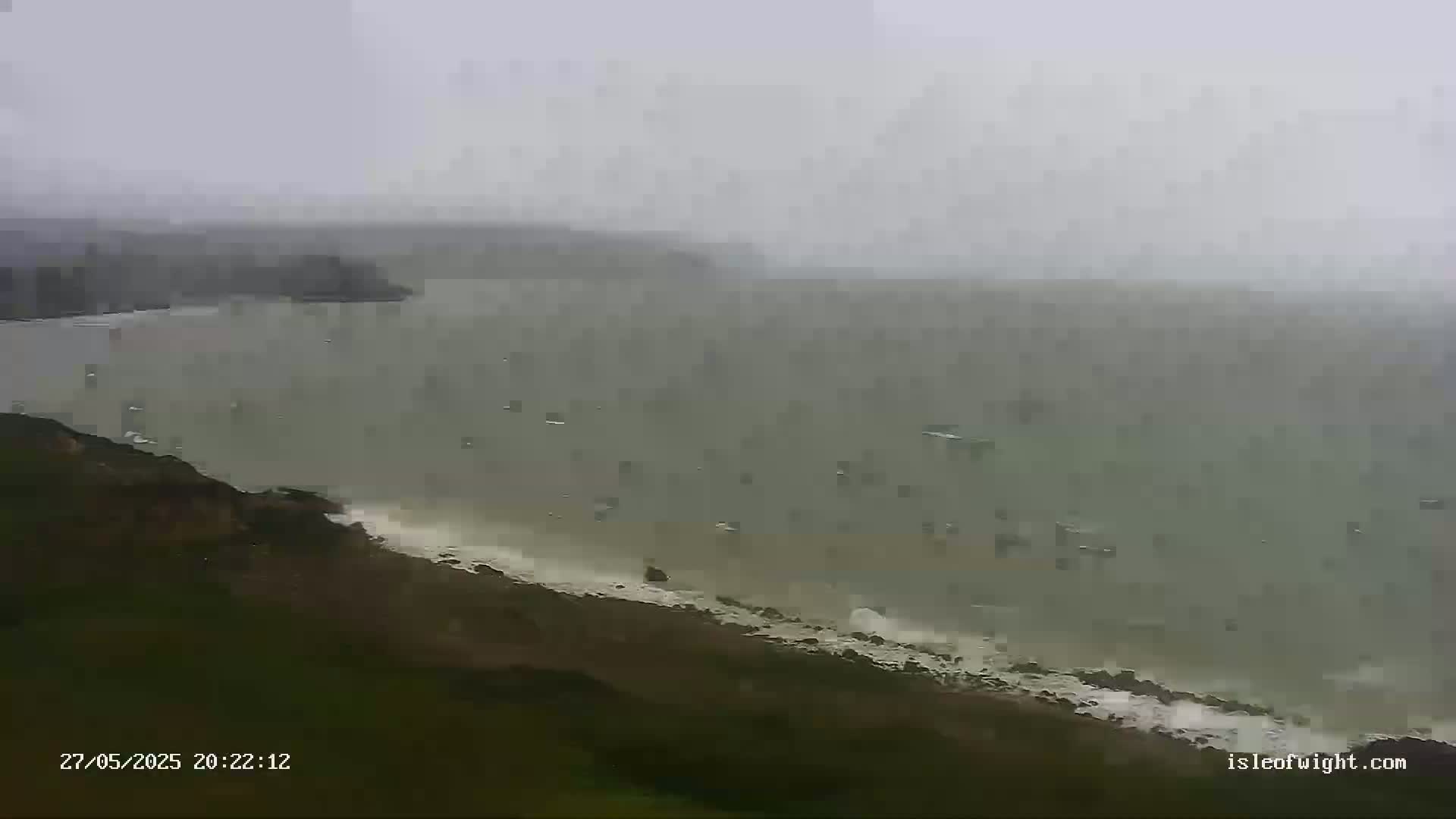 A hazy, overcast day shows a rocky coastline with waves crashing against the shore and a calm body of water extending to a distant landmass.