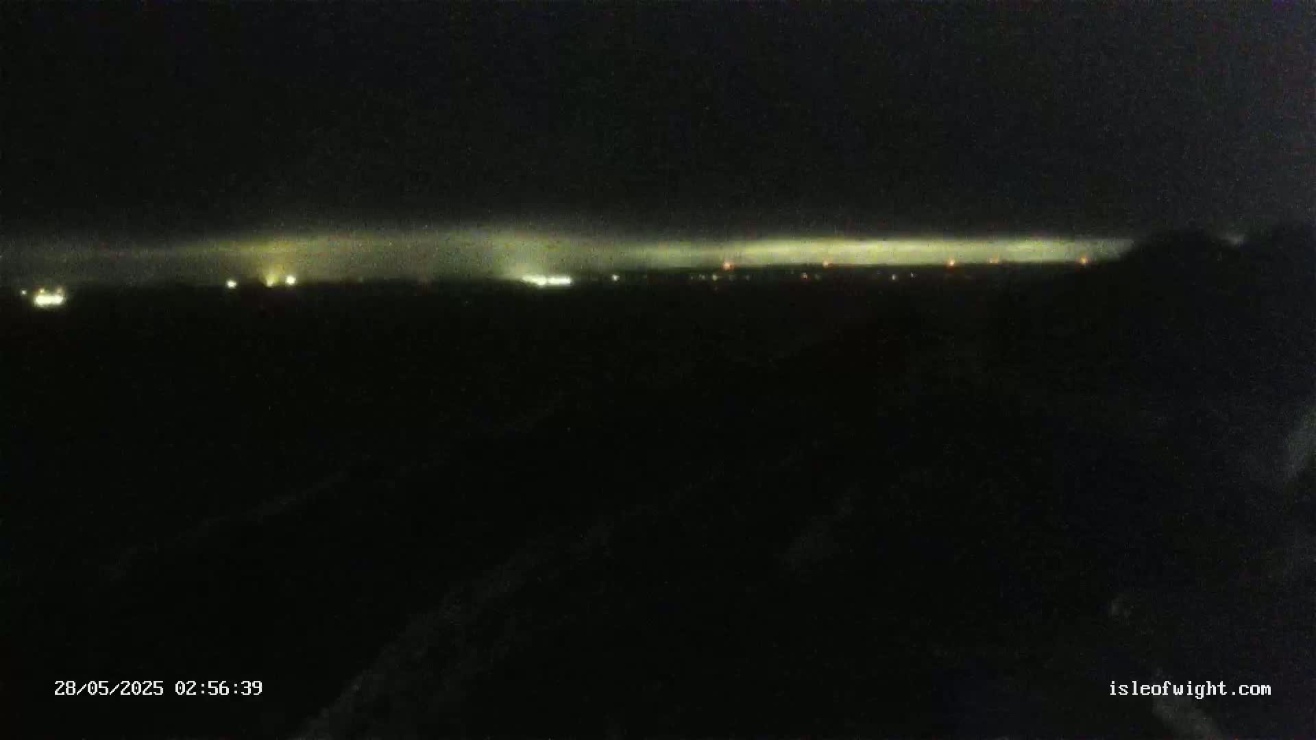 A dark, nighttime landscape shows a distant line of faint lights under a cloudy sky.
