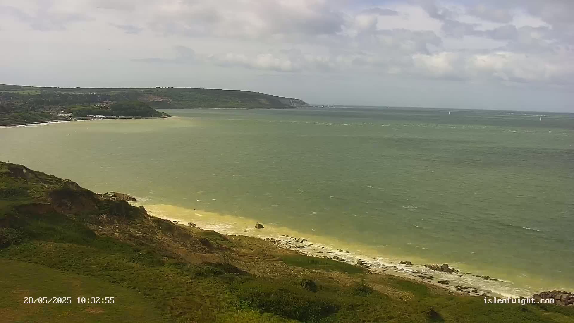 A high-angle view shows a green-tinged ocean under a cloudy sky, with a grassy cliff in the foreground and a distant coastline visible.