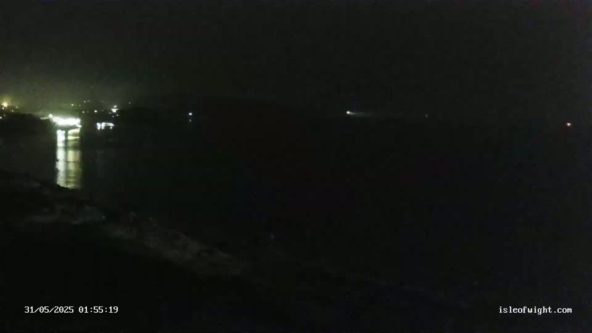 A dark nighttime scene shows a body of water reflecting distant lights, with a dark shoreline in the foreground.