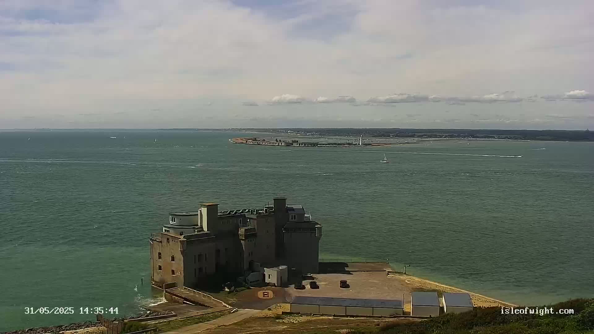 A stone building sits on a coastal point overlooking a calm, greenish-blue sea under a partly cloudy sky.