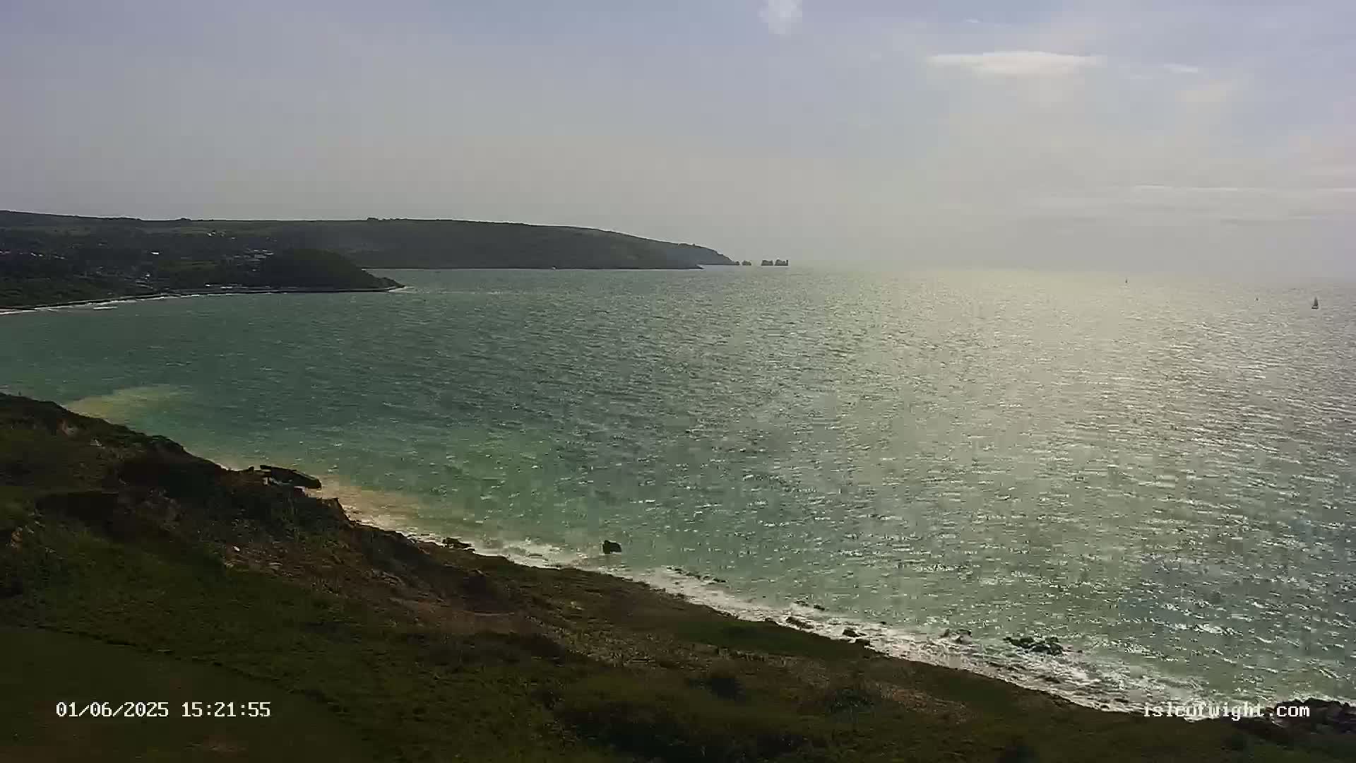 A view from a grassy cliff overlooking a calm, greenish sea with a distant headland and a few small boats under a mostly clear sky.