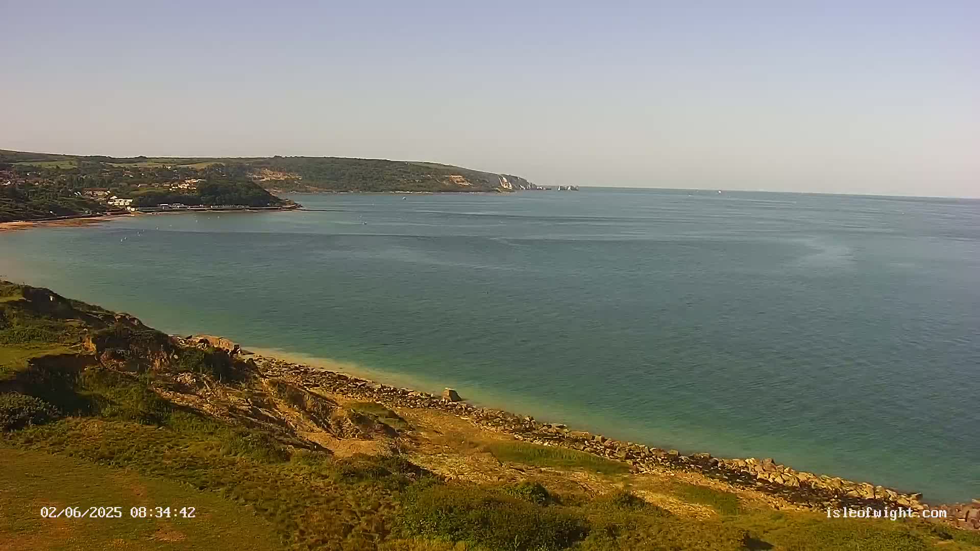 A coastal scene shows a calm, teal sea meeting a sandy beach backed by grassy cliffs and a distant, verdant coastline under a clear sky.