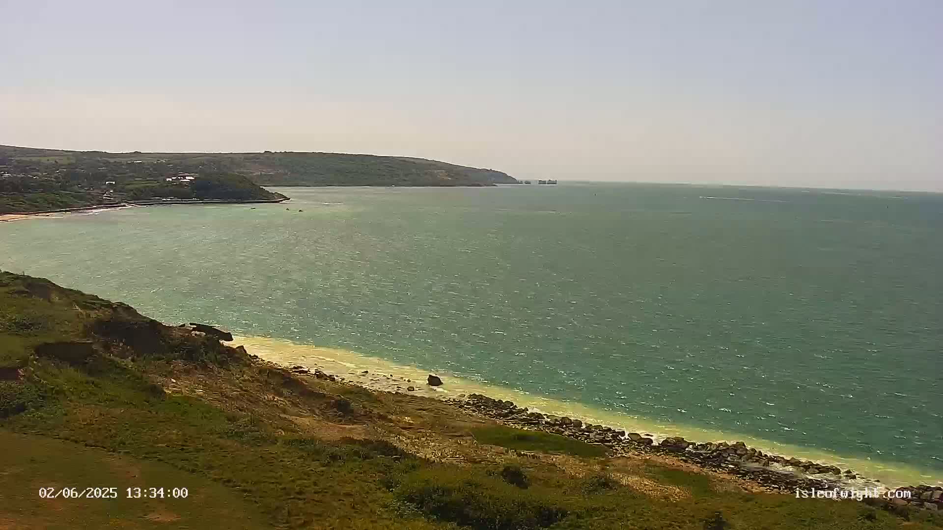 A gently sloping grassy cliff overlooks a calm, greenish-blue sea with a sandy beach; the far shore is visible on a clear day.