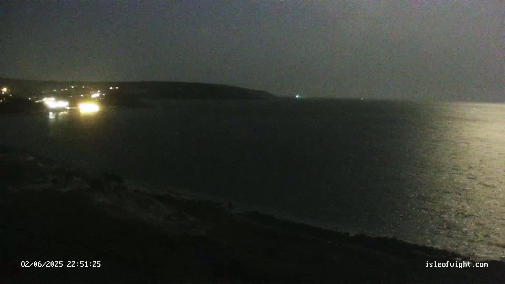 A nighttime view of a dark body of water reflecting light, with a dimly lit coastline in the distance under a dark sky.