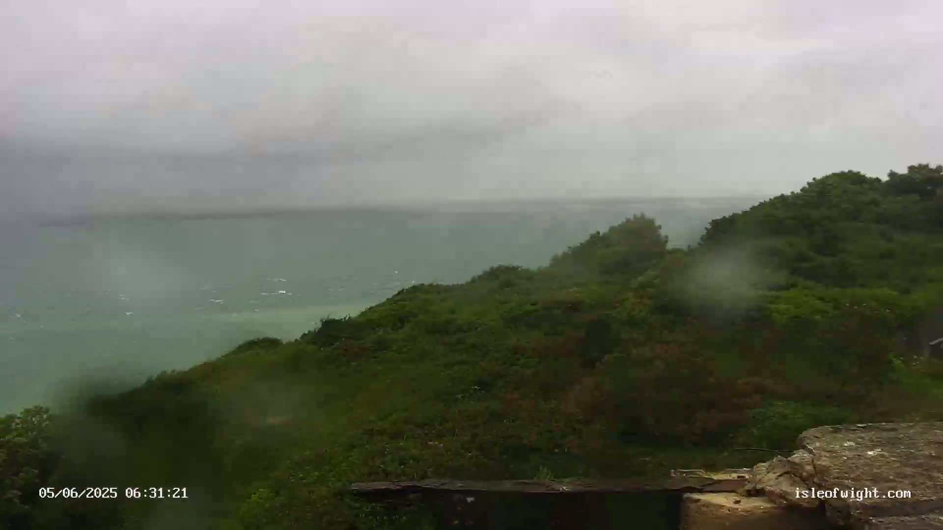 A rain-swept view shows a green, hilly landscape overlooking a choppy sea under a cloudy sky.