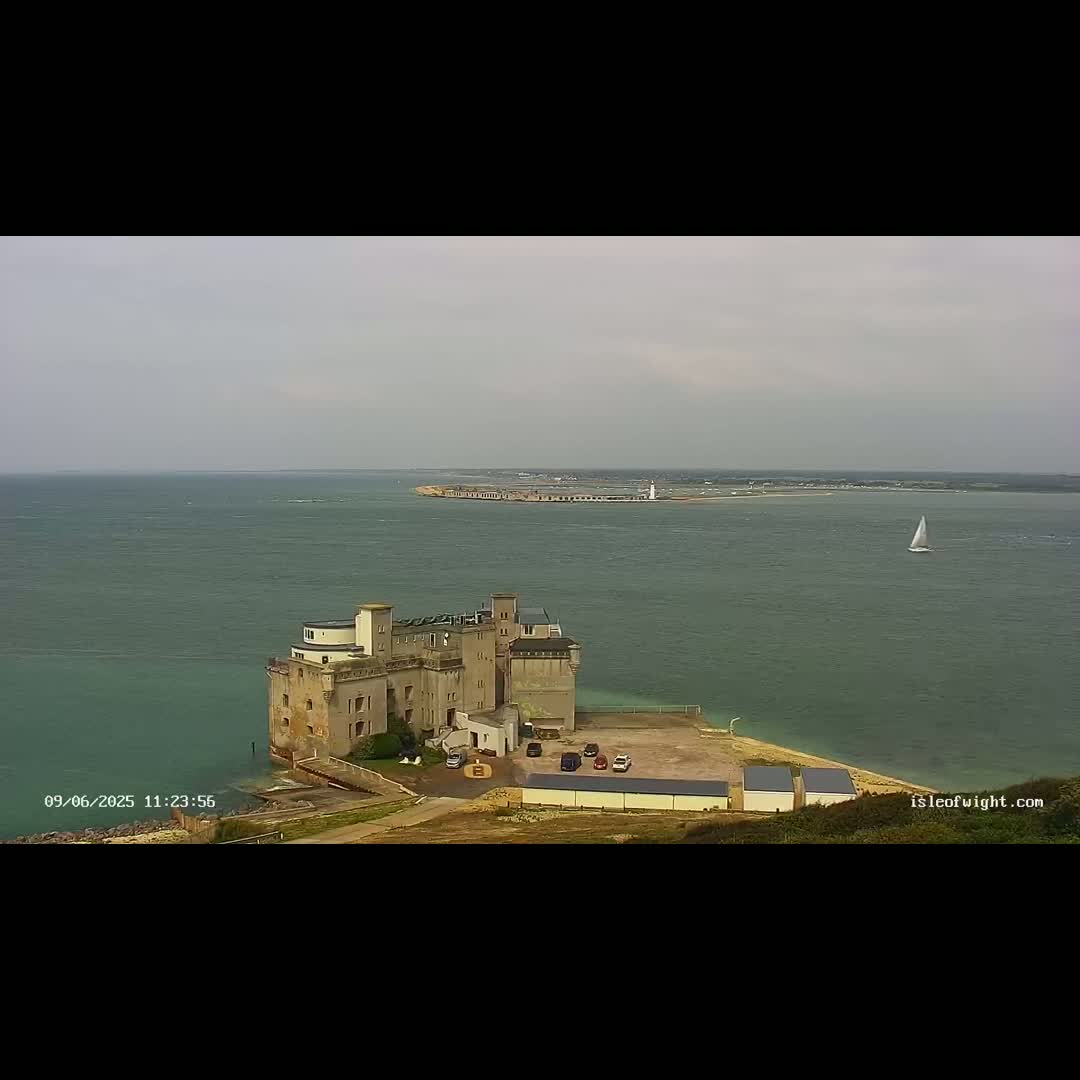 An aerial view shows a stone castle-like structure on a coast overlooking a calm body of water with a distant landmass and a sailboat.