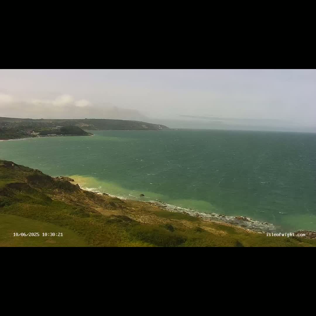 A windy day shows a view of a green sea meeting a grassy cliff overlooking a sandy beach.