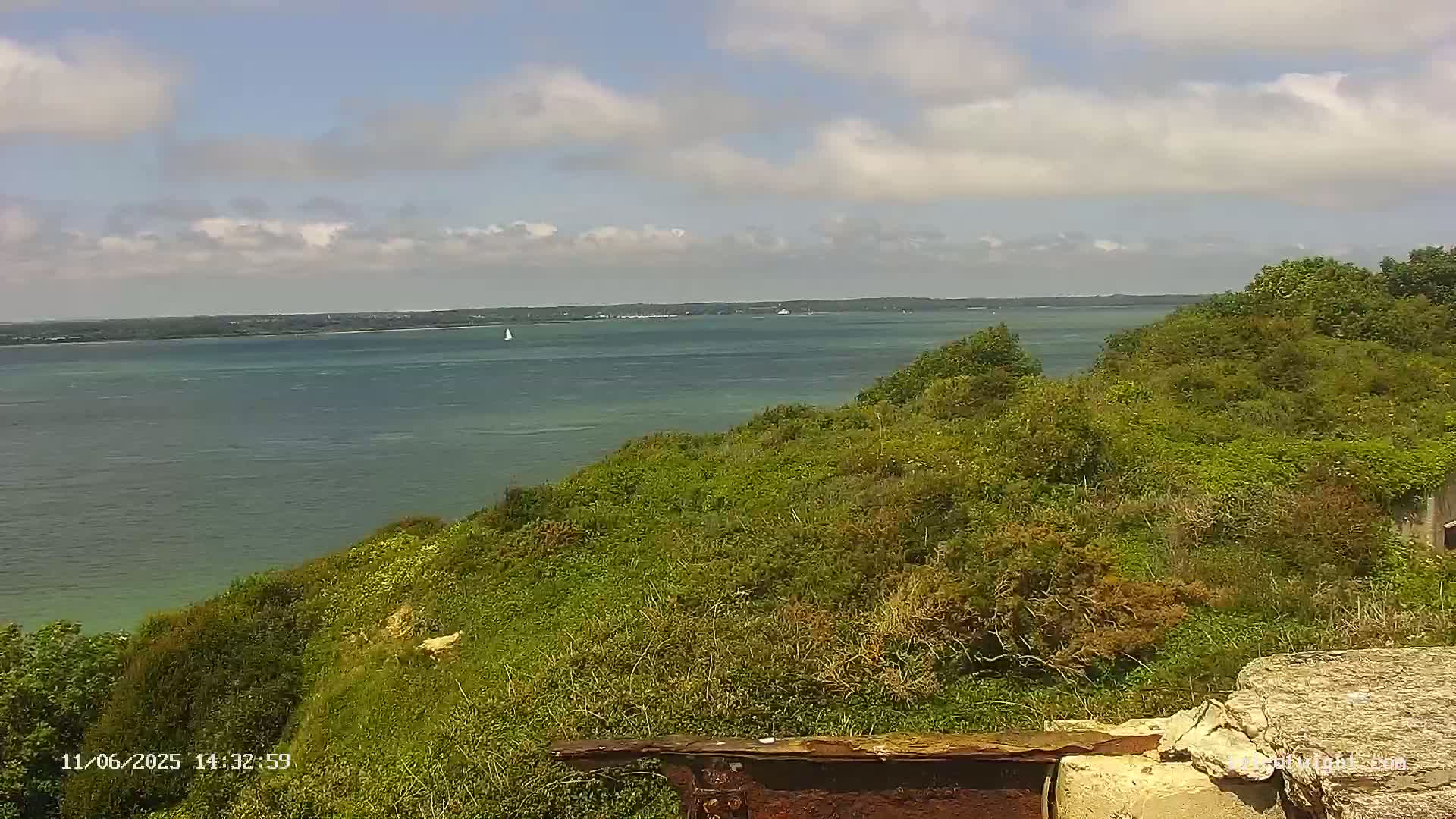 A partly cloudy day overlooks a calm body of water from a verdant, hilly shoreline with some remnants of a structure in the foreground.