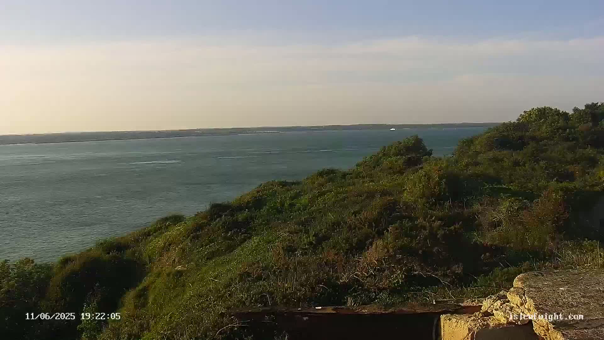 A calm body of water is viewed from a verdant clifftop on a partly sunny day.