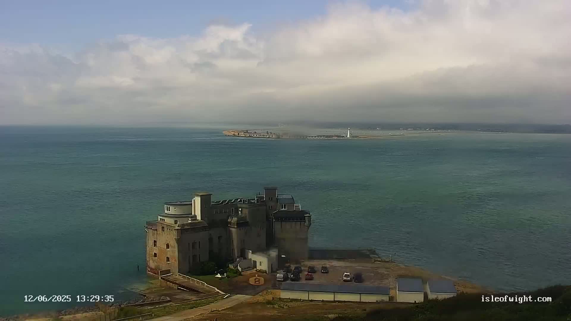 A large stone building sits on a coastal cliff overlooking a calm, overcast sea with a distant landmass visible on the horizon.