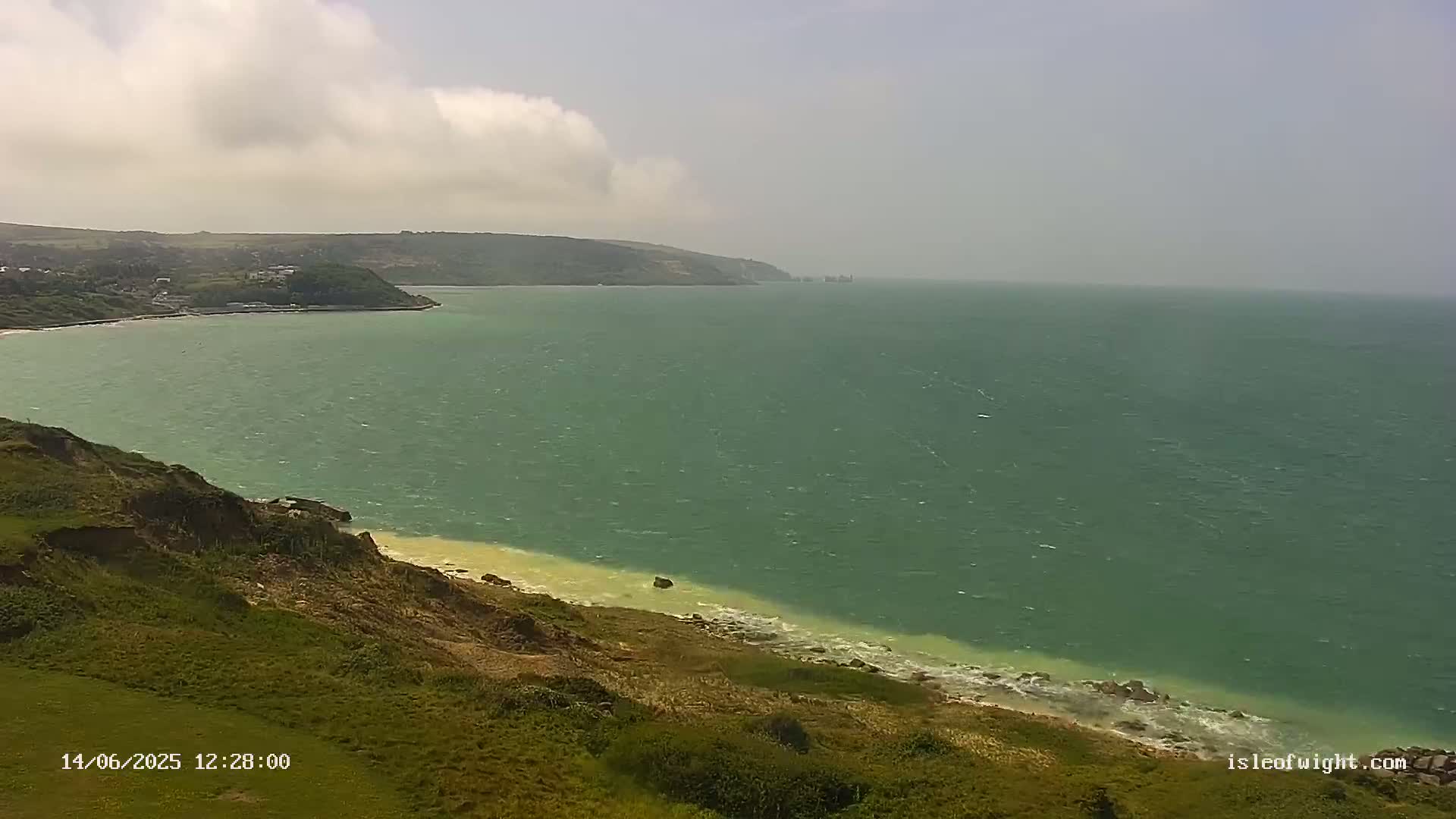 A green sea with small waves laps a rocky shore beneath a partly cloudy sky.