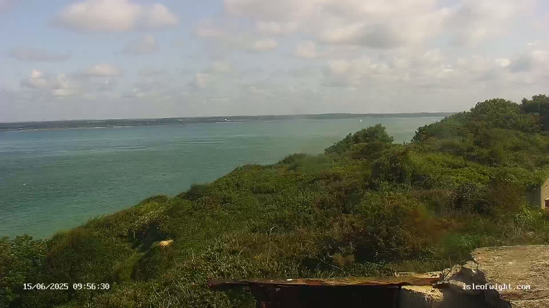 A partly cloudy day shows a calm, green body of water extending to a distant landmass, viewed from a high vantage point above a lush, green hillside.