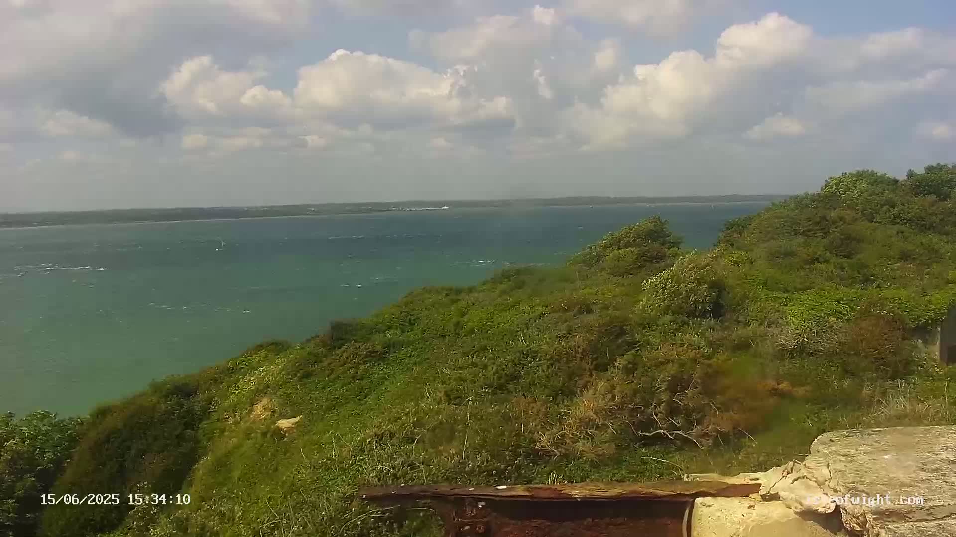 A partly cloudy day reveals a view of a calm, green body of water from a verdant clifftop.