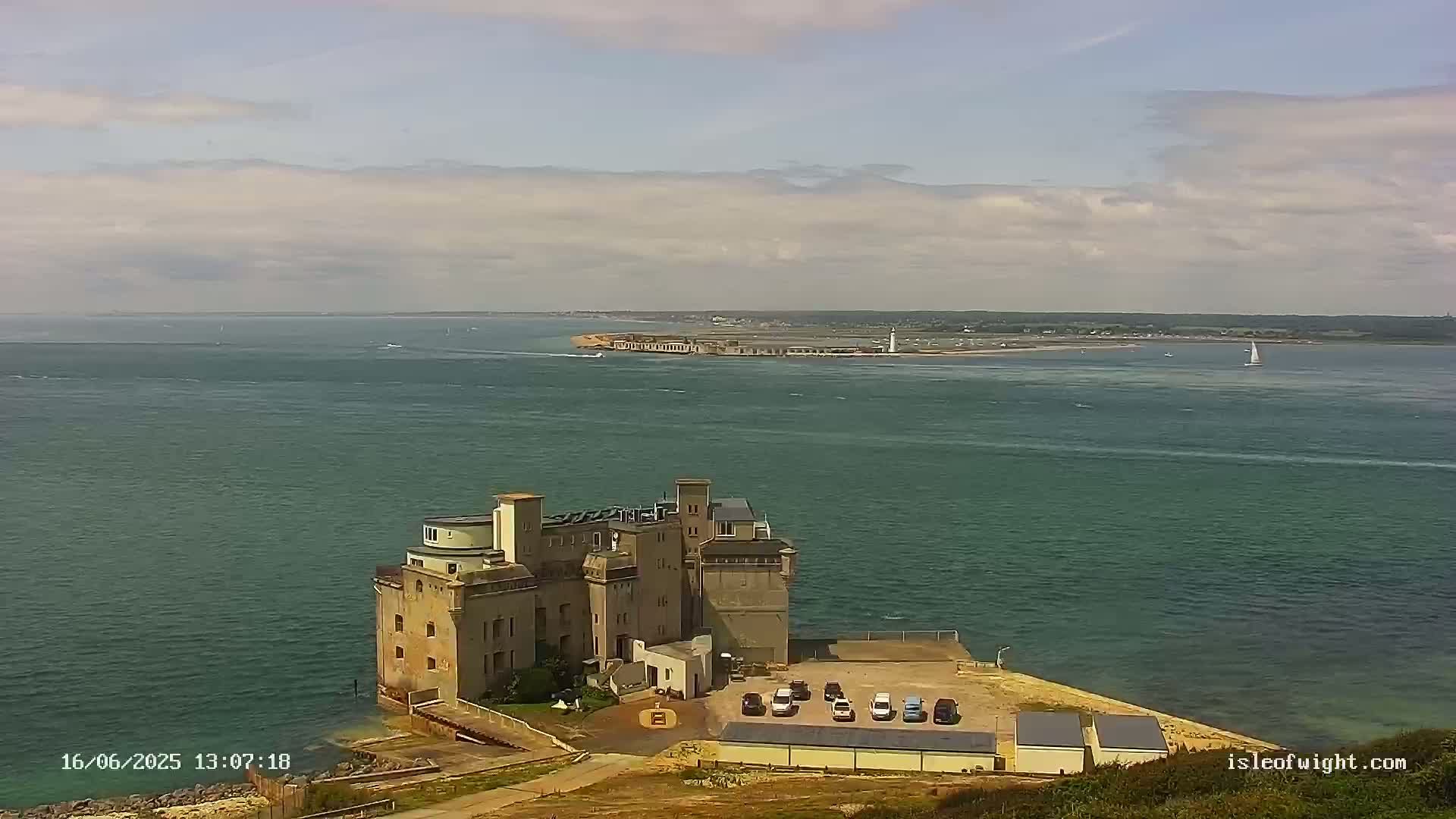 A stone building resembling a castle sits on a coastal point overlooking a calm body of water under a partly cloudy sky, with a distant landmass visible in the background.
