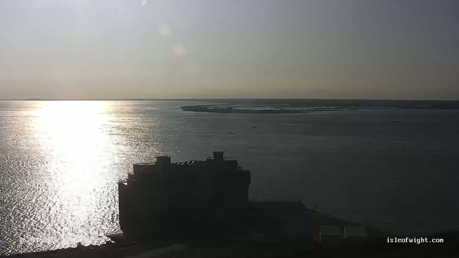A sunlit body of water stretches to a distant shoreline under a mostly clear sky, with a dark building silhouetted in the foreground.