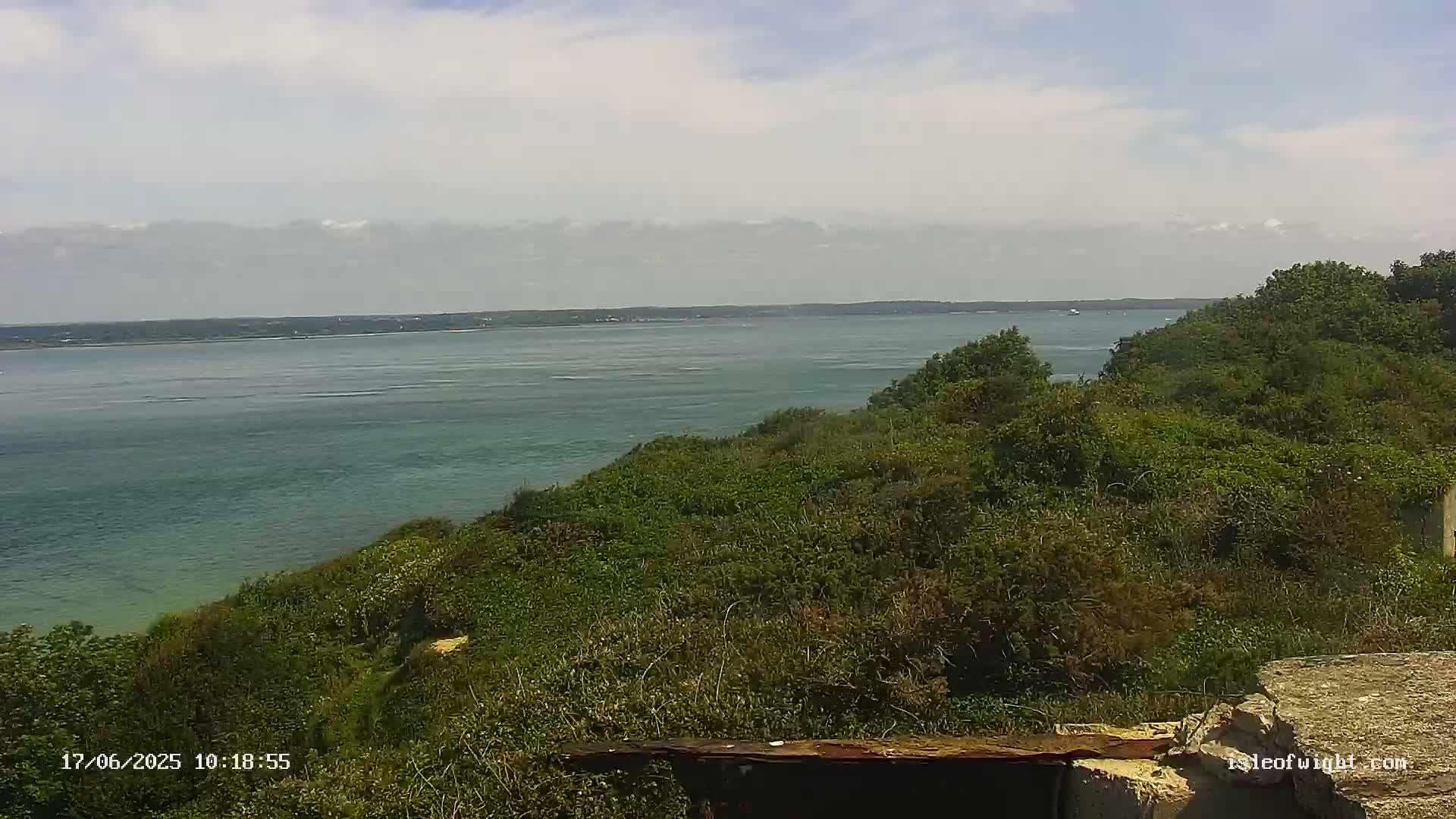 A partly cloudy day overlooks a calm body of water from a verdant clifftop, with remnants of a structure in the foreground.