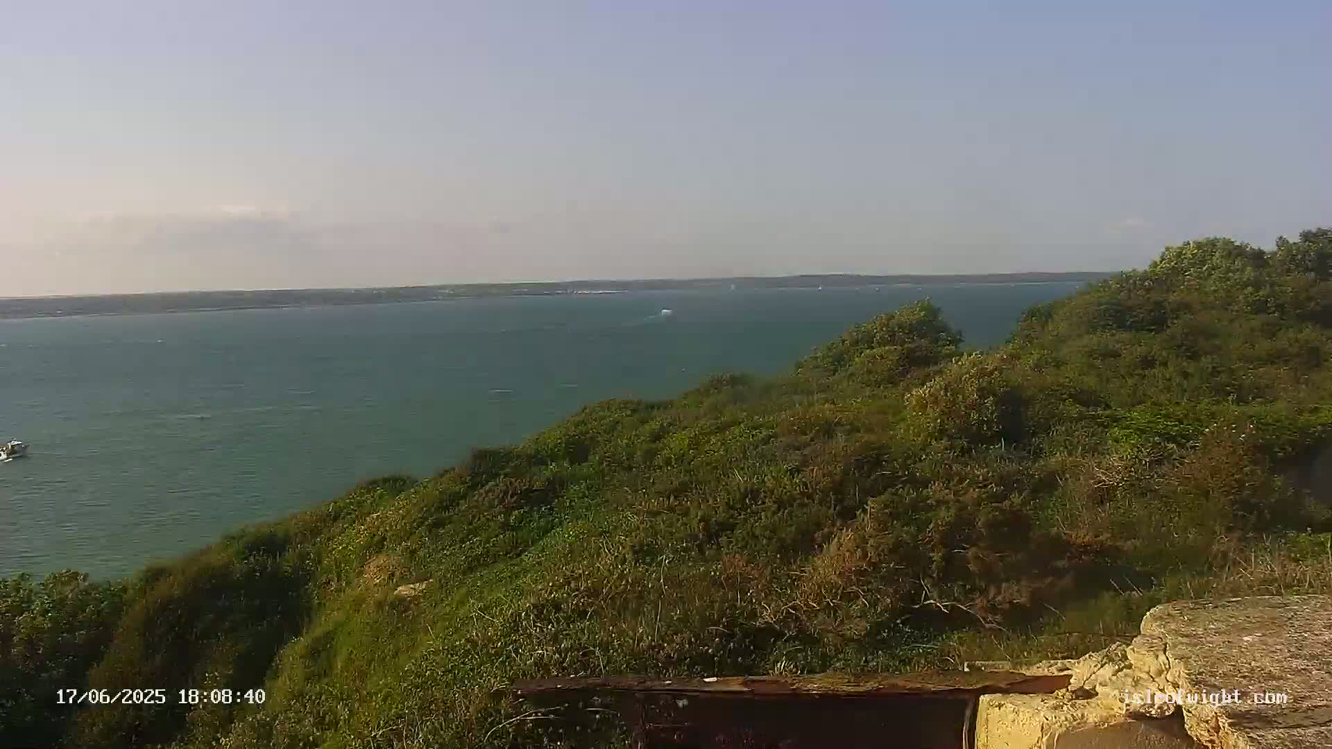 A calm, expansive body of water is visible from a verdant clifftop, with a few boats in the distance under a mostly clear sky.