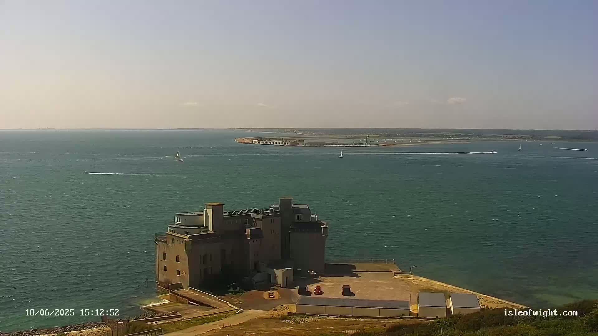 A large stone building sits on a point of land overlooking a calm, green body of water with several small boats visible in the distance on a sunny day.