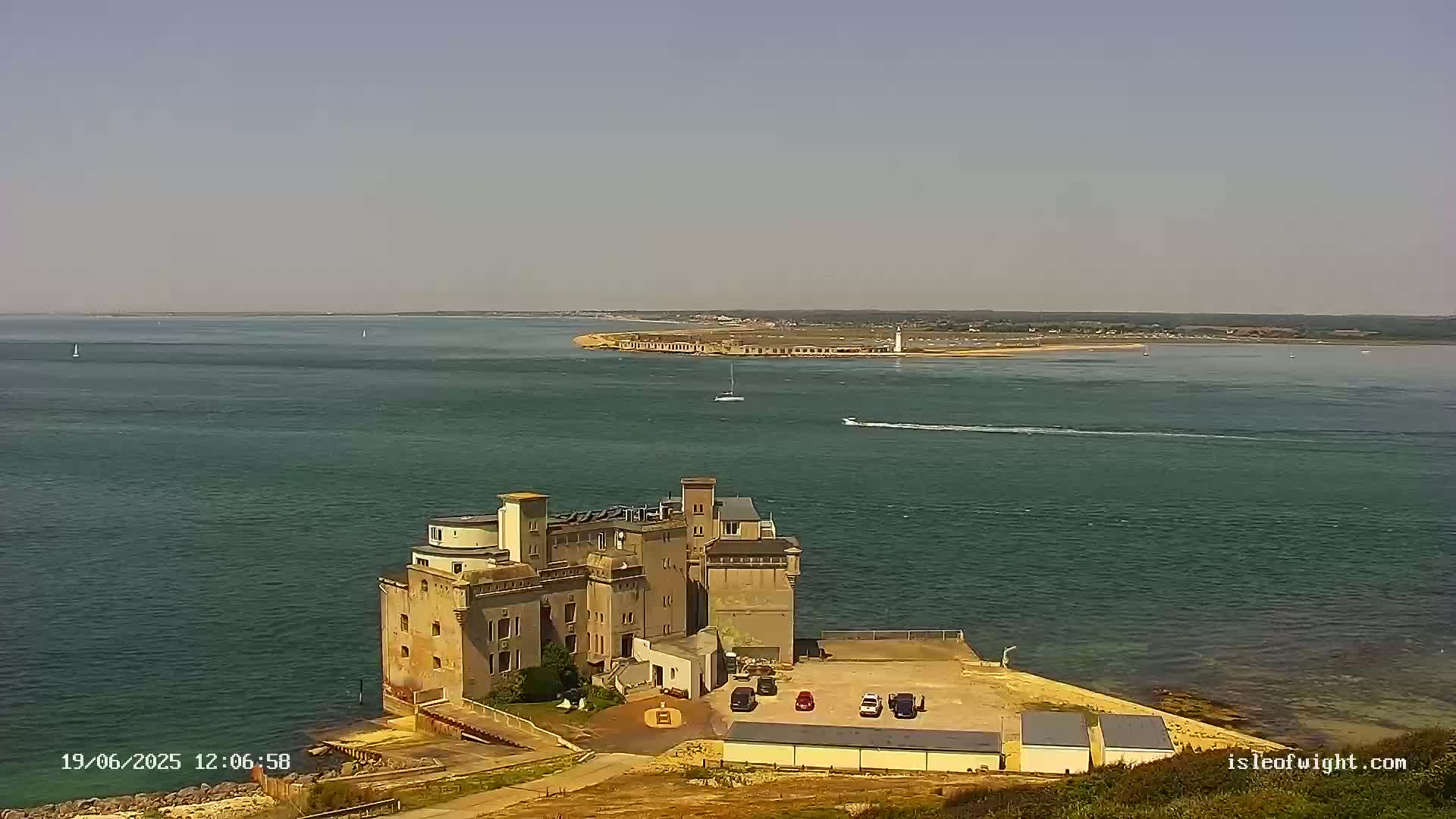 A large stone building sits on a peninsula overlooking a calm body of water on a sunny day, with another landmass visible in the distance.