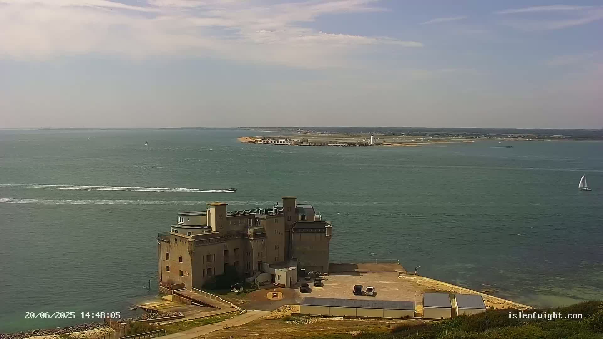 A high-angle view shows a stone building situated on a point of land jutting into a calm body of water, with a distant shoreline and several small boats visible on a partly sunny day.