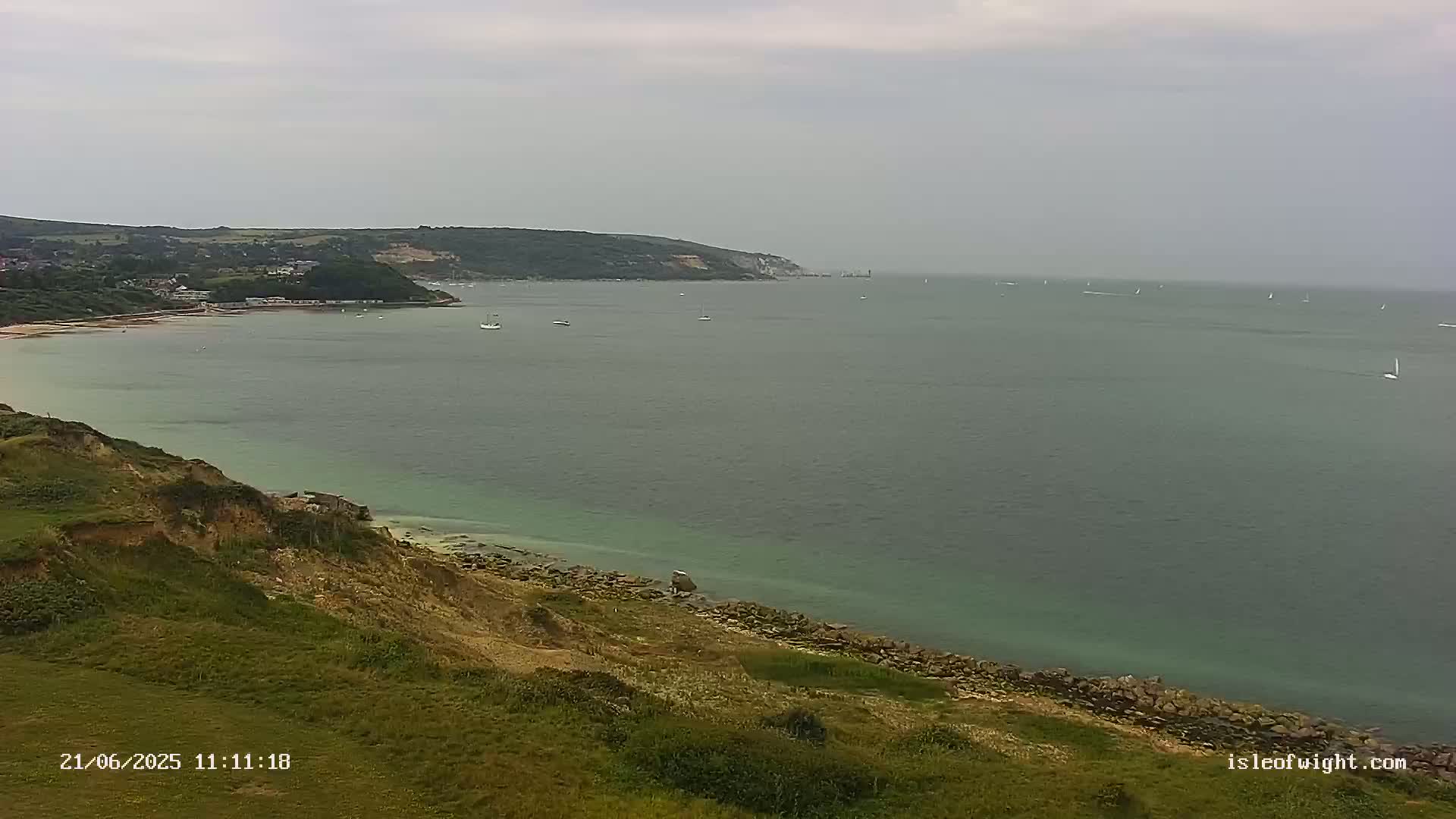 A grassy cliff overlooks a calm, greenish-grey sea with several small boats and a distant coastline under an overcast sky.