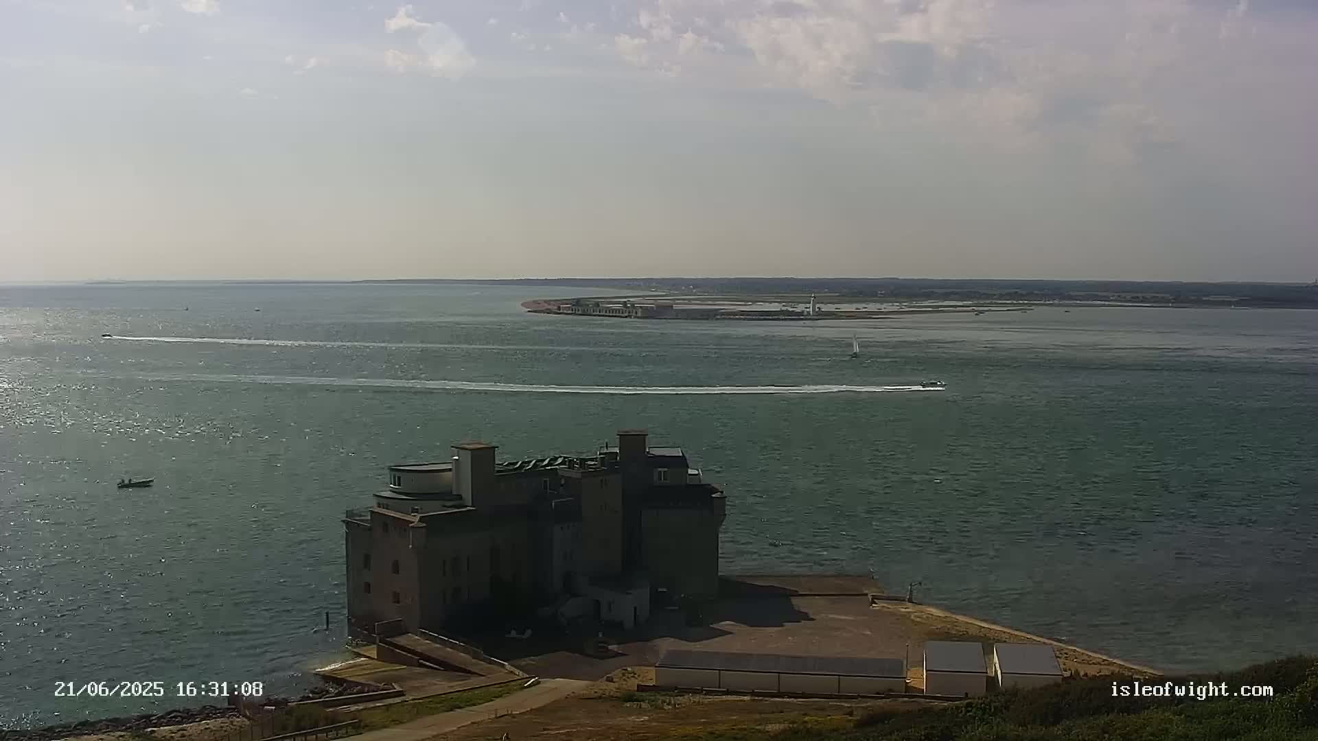 A multi-storied stone building sits on a point of land overlooking a calm body of water with several small boats, under a partly cloudy sky.