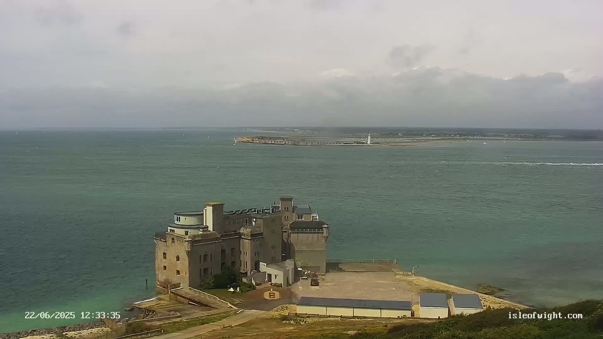 A large, stone building sits on a coastal point overlooking a calm, greenish-grey sea under a cloudy sky.