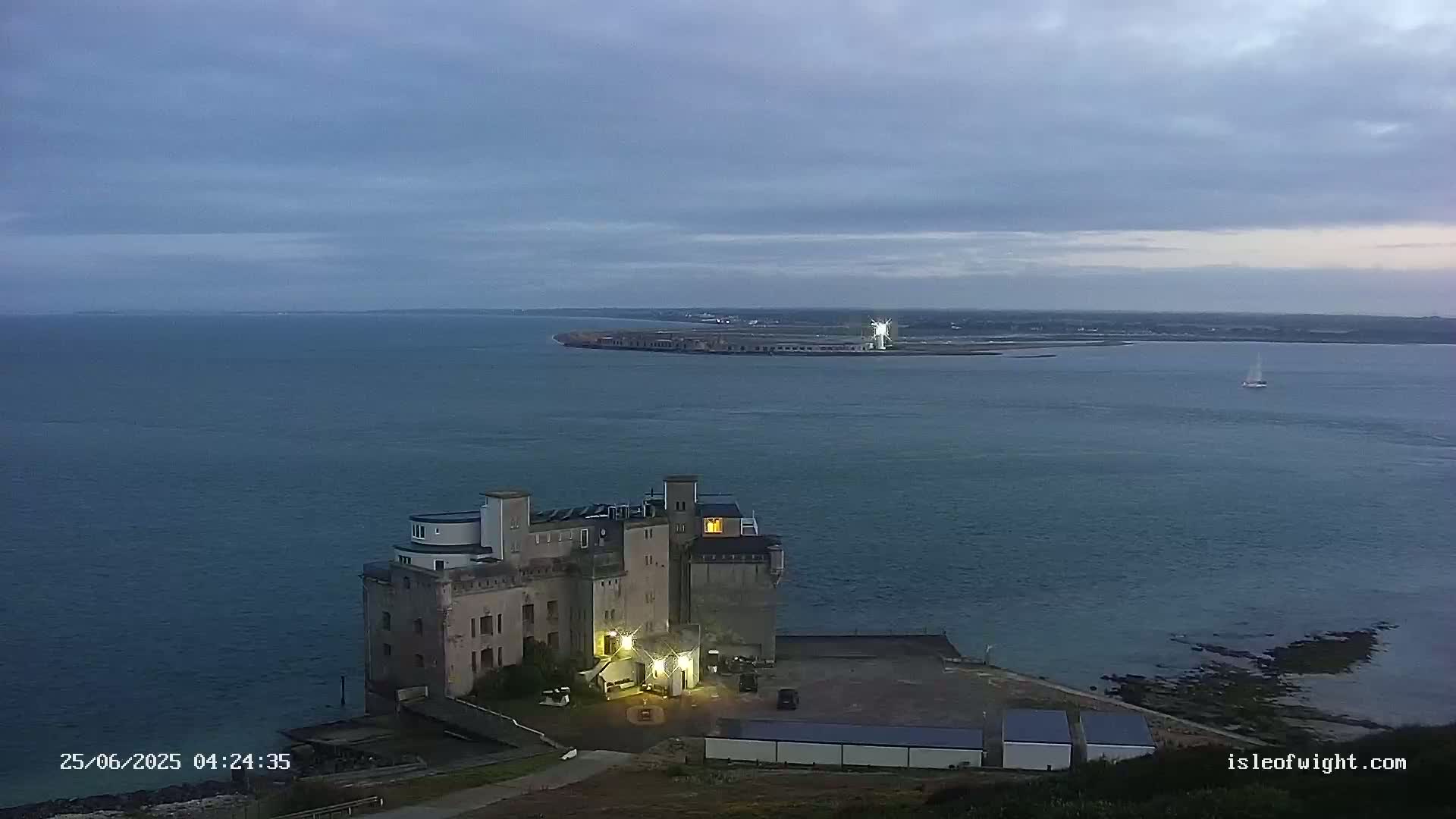 A large, castle-like building with lights on sits on a rocky shoreline overlooking calm, dark water under an overcast sky, with a distant ship and a sailboat visible on the water.