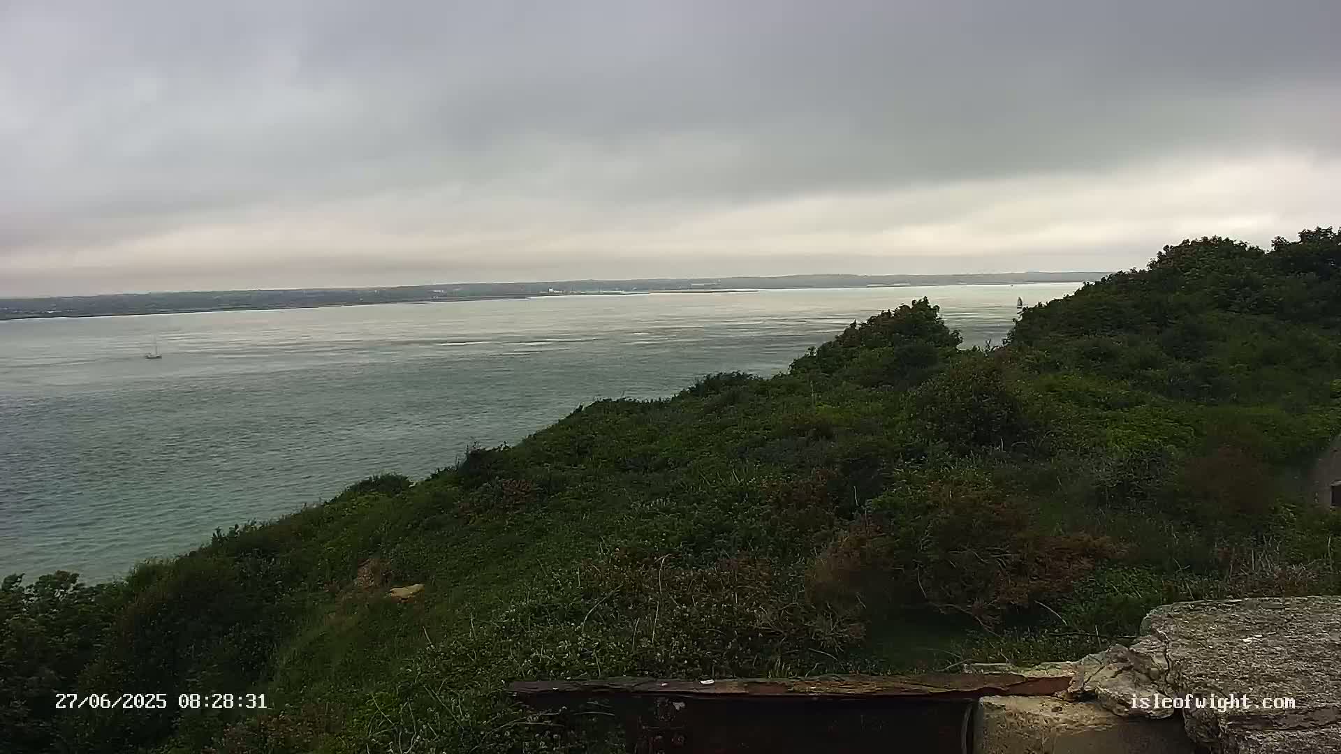 A view from a green hill overlooking a calm, grey sea under an overcast sky.