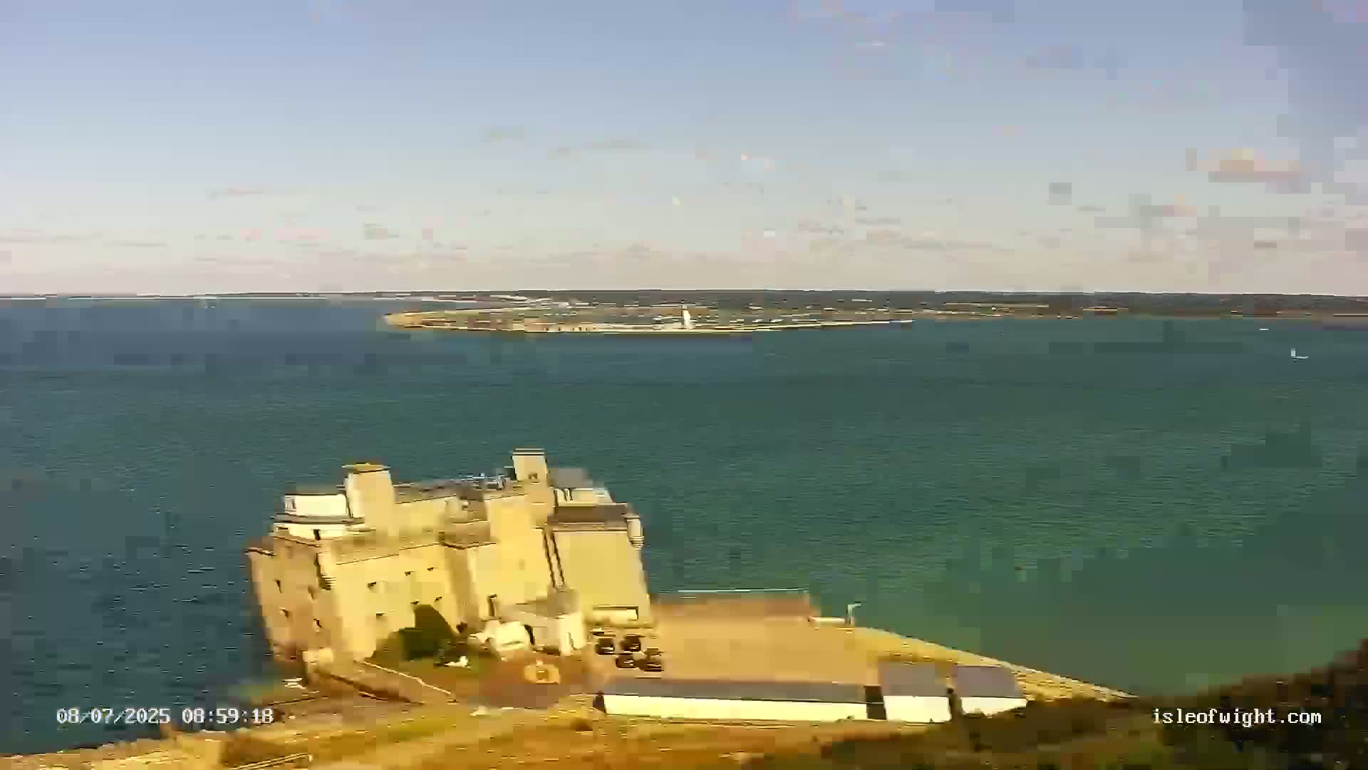 A large stone building sits on a cliff overlooking a calm, teal ocean, with land visible in the distance under a mostly clear sky.