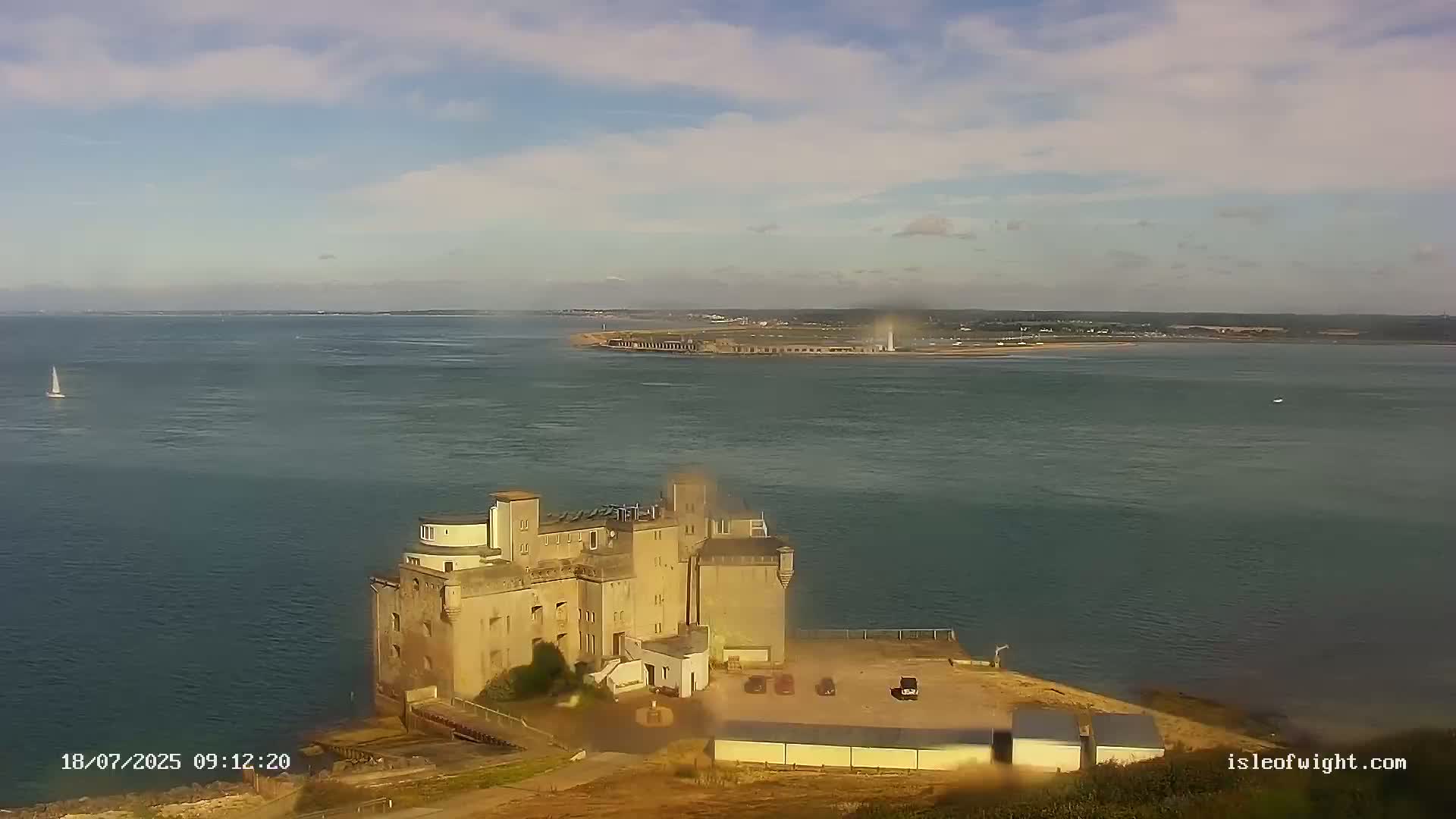 A large stone building sits on a small peninsula overlooking a calm body of water under a partly cloudy sky.