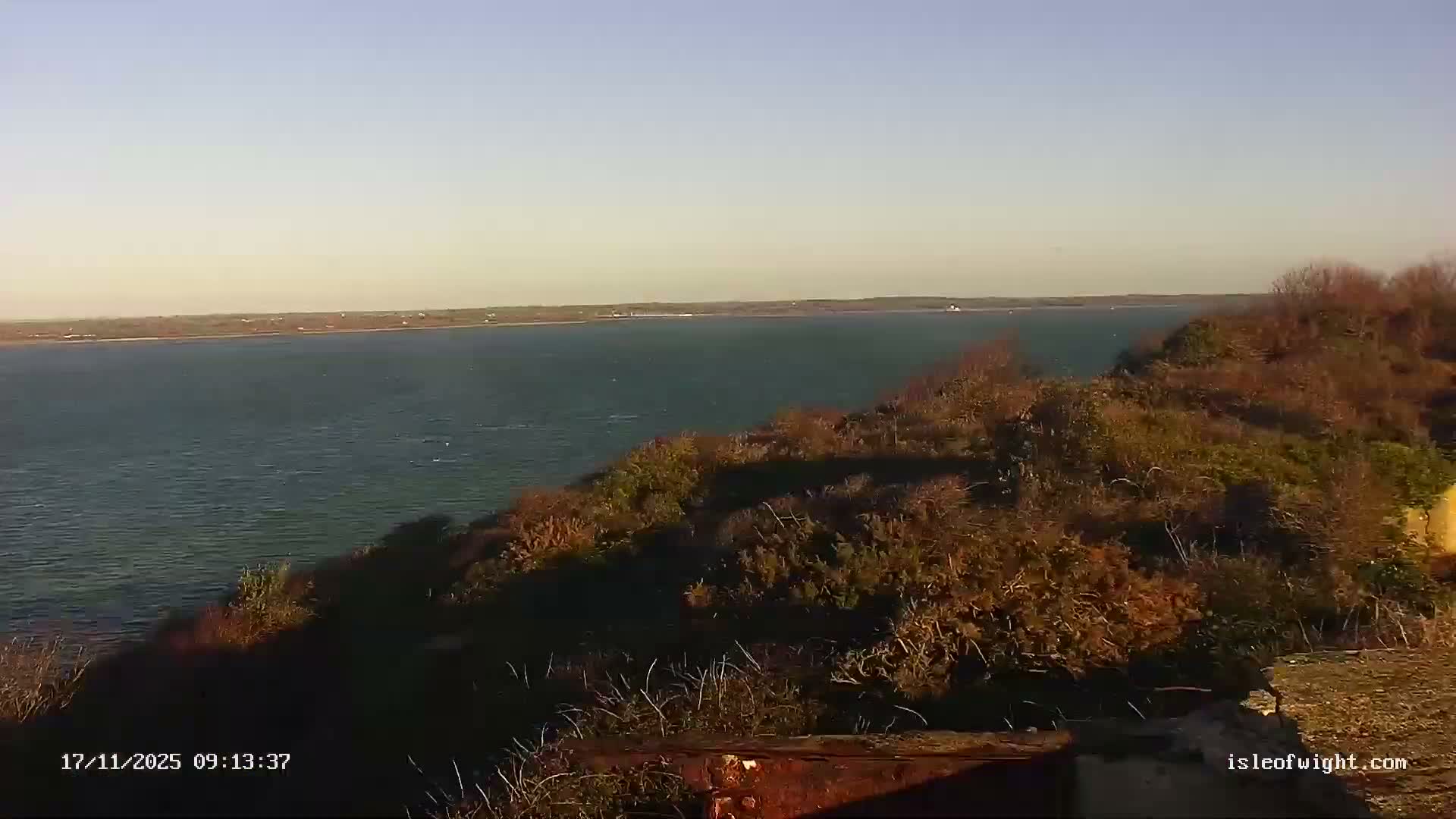 The image shows a wide body of water bordered by a distant shoreline and a brush-covered hillside in the foreground, under a clear, sunny sky.
