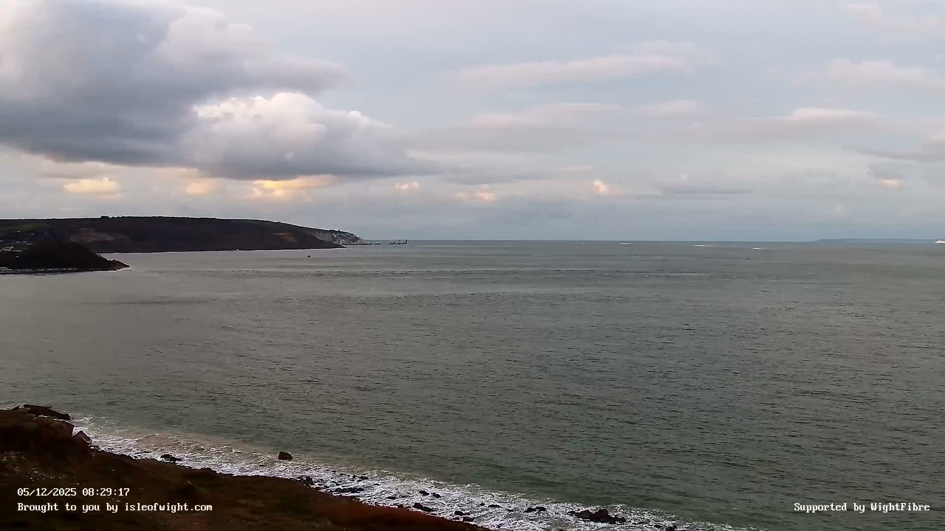 An overcast morning scene reveals choppy grey-green coastal waters, a rocky shore, and dark distant cliffs under a cloudy sky with faint golden light peeking through.