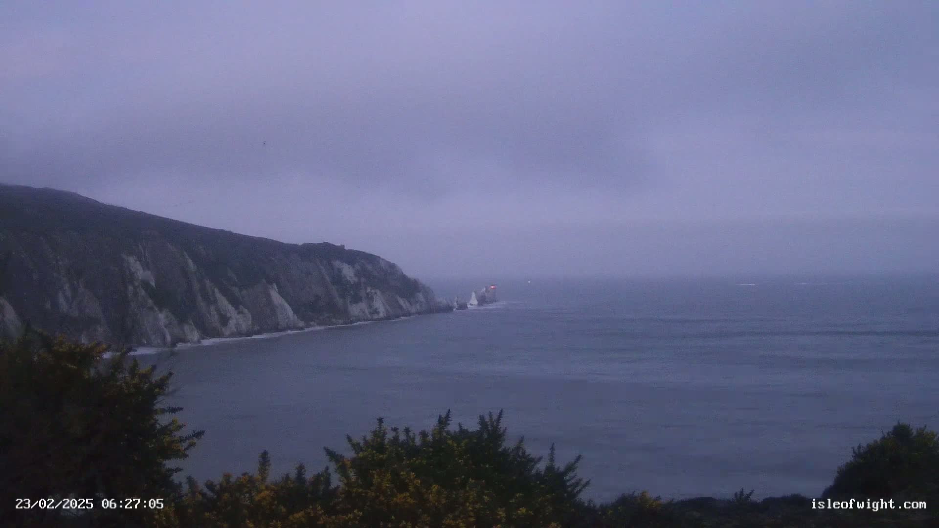 A hazy, overcast day reveals a steep, cliff-lined coast meeting a calm, gray sea with a distant vessel.