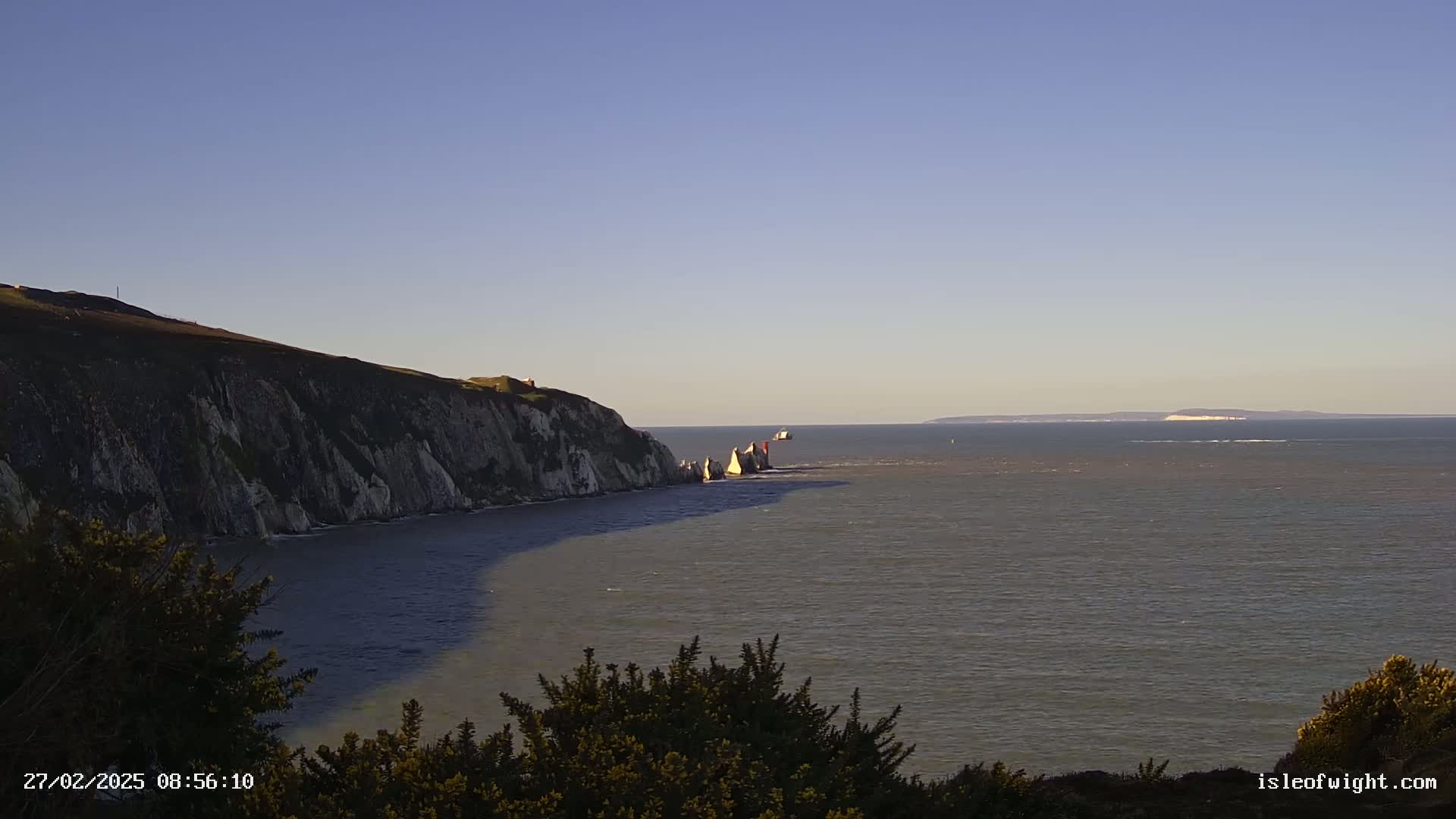A coastal scene under a clear blue sky shows white cliffs, several rock formations in the water, and a distant landmass on the horizon.