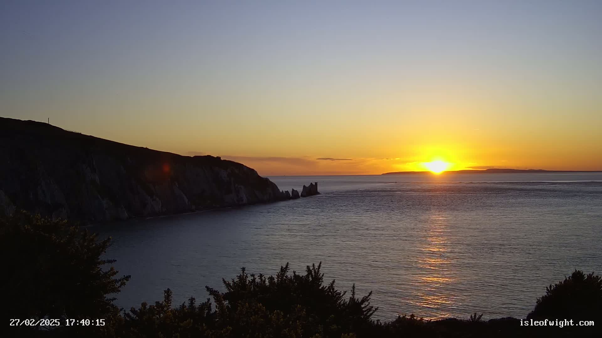 A vibrant sunset over a calm ocean illuminates a dark cliff and distant rock formations.