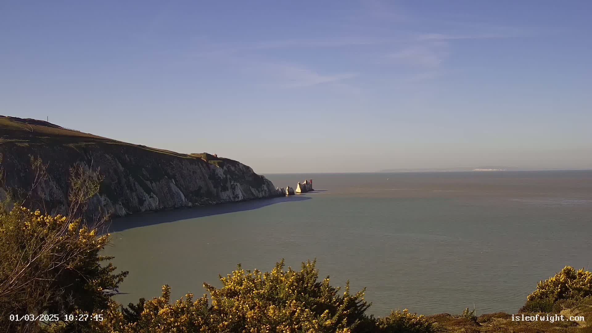 A coastal view shows a high white cliff overlooking a calm sea with a smaller rock formation in the distance under a clear blue sky.