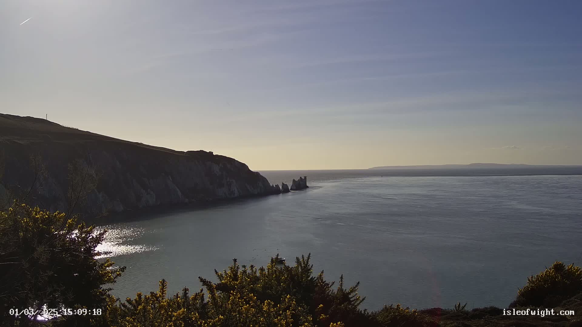 A bright, sunny day reveals a coastal scene with a dramatic cliff face overlooking a calm sea, where a small group of distant rocks are visible.
