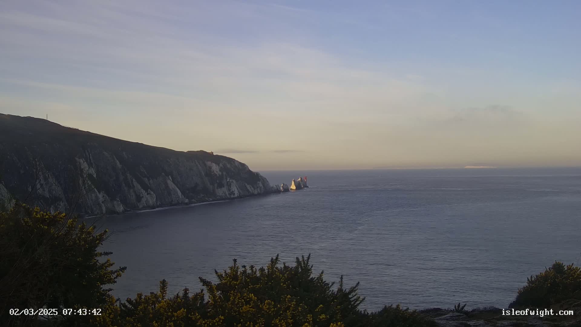 A coastal scene at sunrise shows a cliff face meeting a calm ocean with a small group of rocky islands in the distance.