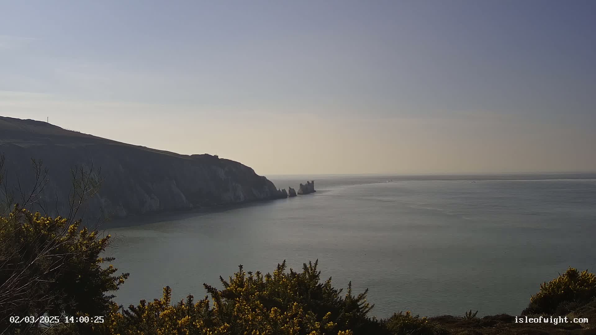A coastal view shows a calm sea extending to a hazy horizon, with a high, dark cliff face to the left, and a smaller rock formation further out in the water.