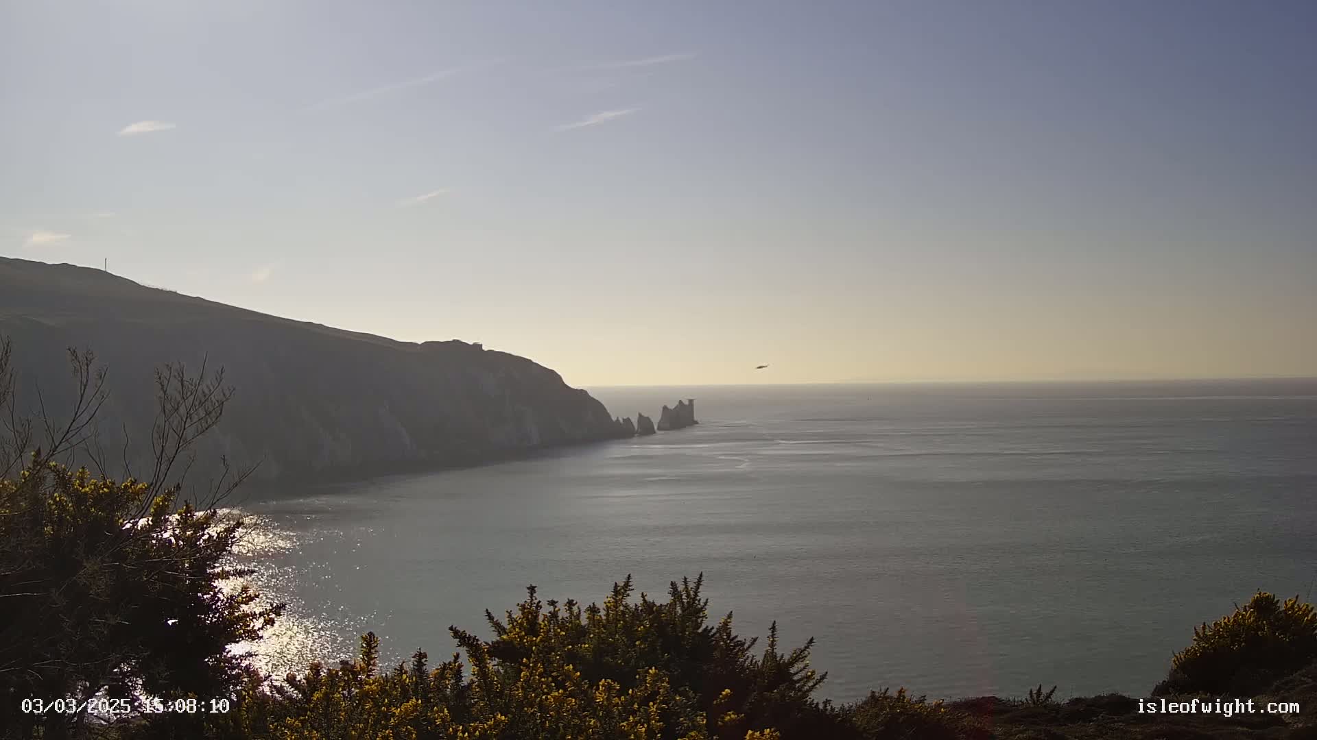 A coastal scene under a bright, clear sky shows a cliffside, a calm sea, and distant rock formations.